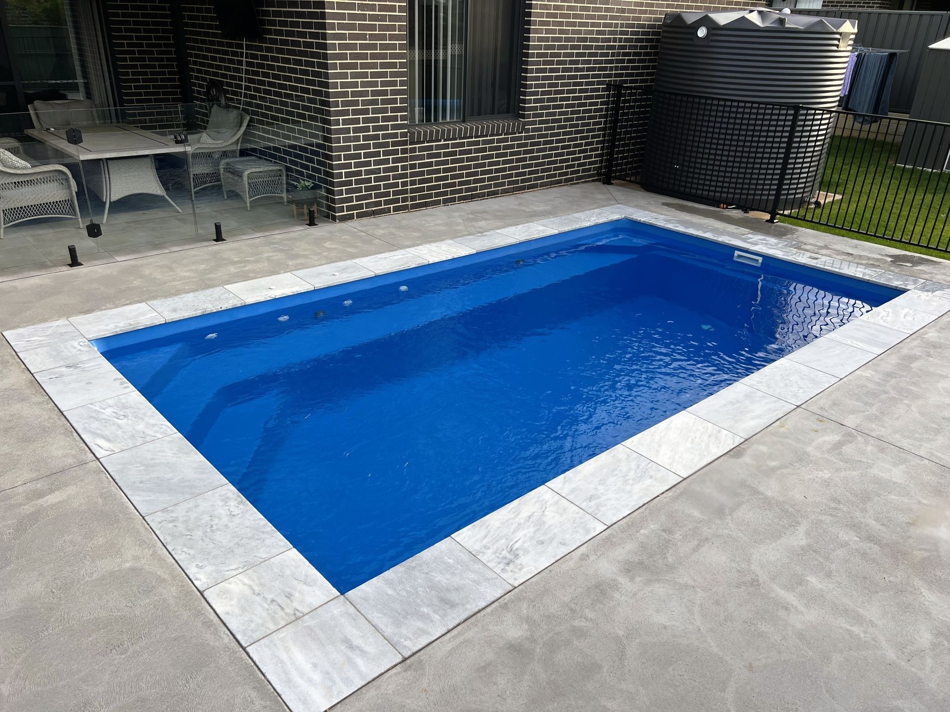 Rectangular blue swimming pool with gray tiled border next to a brick building and patio.
