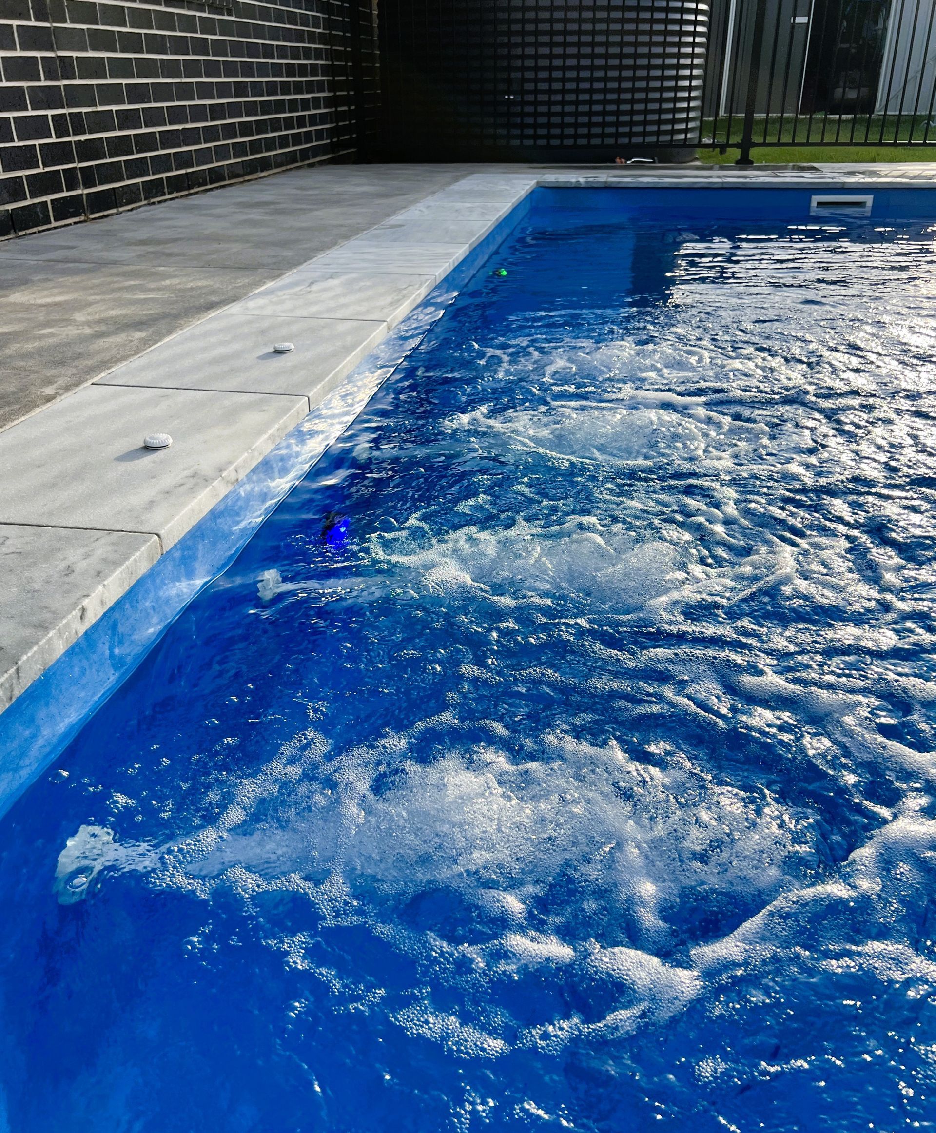 Blue swimming pool with water jets. Concrete border next to a brick wall.