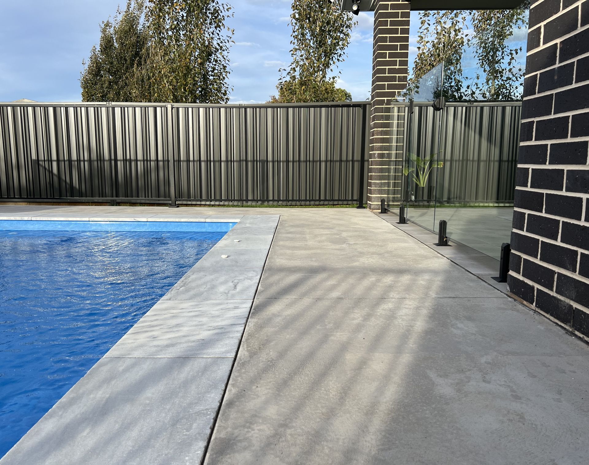 Poolside view: blue pool, grey concrete, patterned fence, brick wall, glass panel.