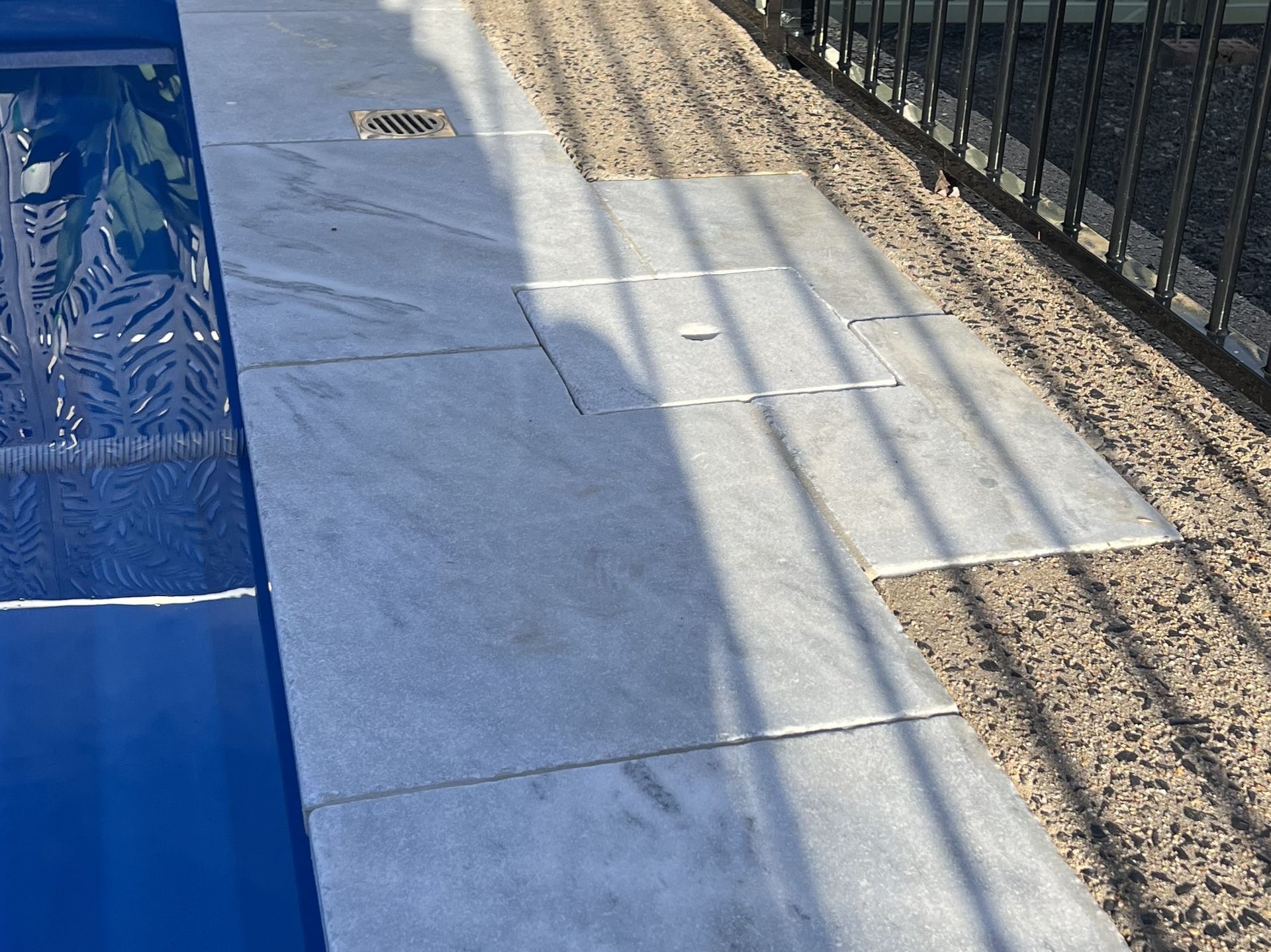 Poolside with gray stone tiles, drain, gravel, and black metal fence. Blue water on left.