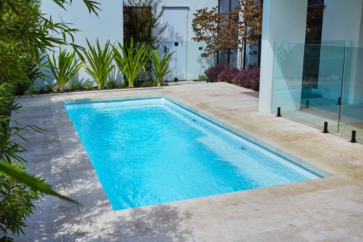 Rectangular pool with clear blue water, bordered by light stone patio and white walls, lush greenery.