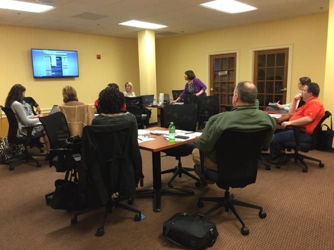 People in a meeting room, some at tables with computers, one standing and speaking. A screen displays content.