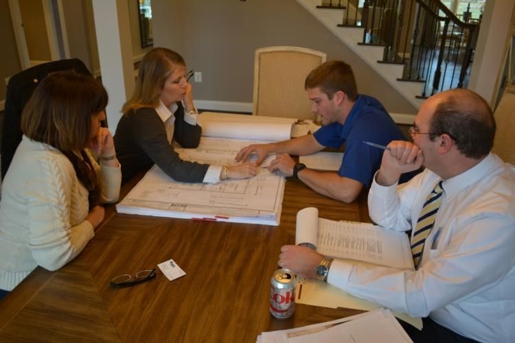 Four people reviewing blueprints around a table. One person points, others look on. Stairs are in background.