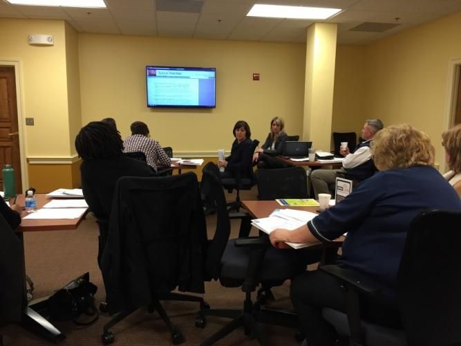 People in a meeting room, facing a screen displaying text. Tables, chairs, and notebooks are visible.