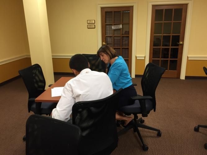 A woman and a person in a white shirt sit at a table looking at a document. They are in a room with a brown carpet.