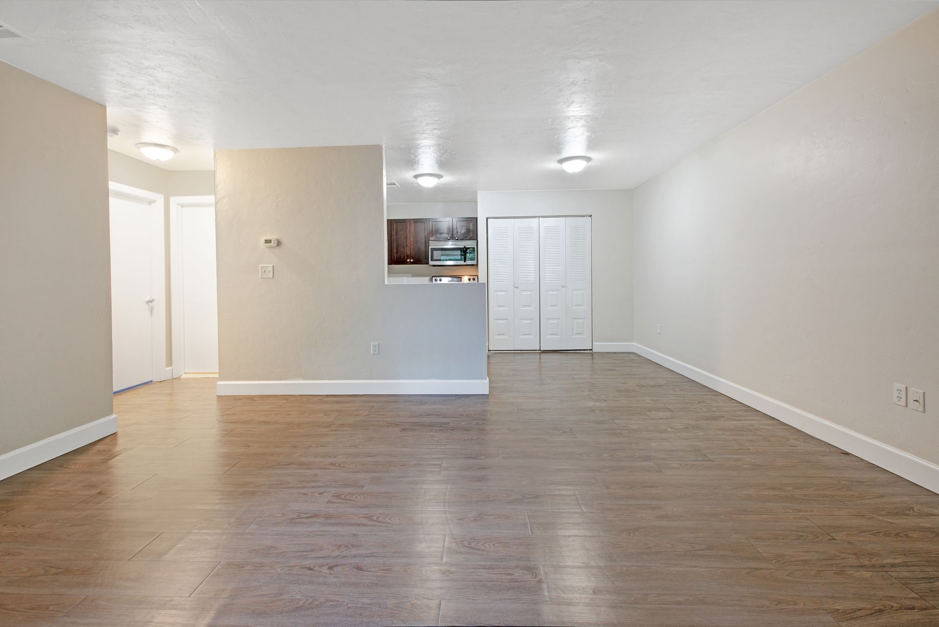 An empty living room with hardwood floors and a kitchen in the background.