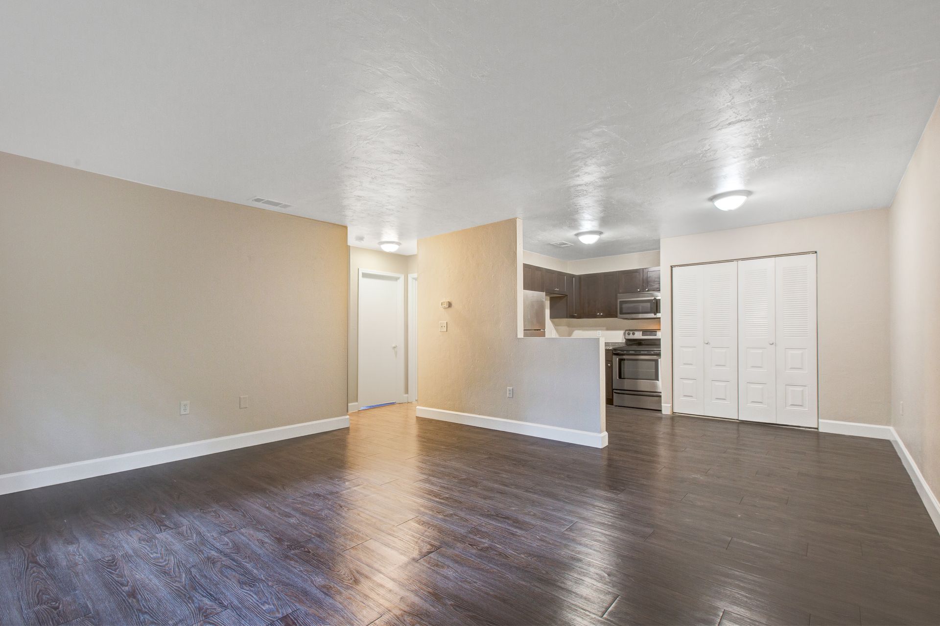 An empty living room with hardwood floors and a kitchen in the background.