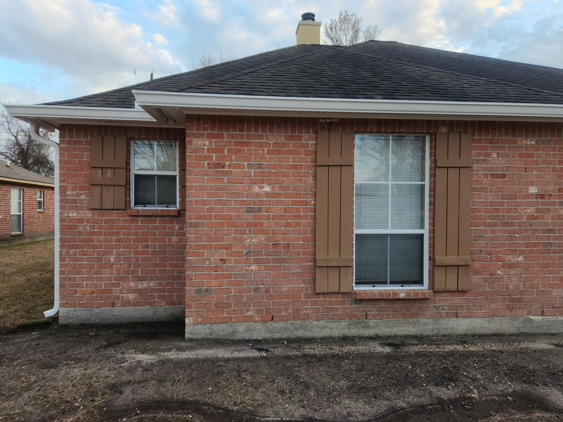 Single-story red brick house exterior with dark shingled roof and two front windows.
