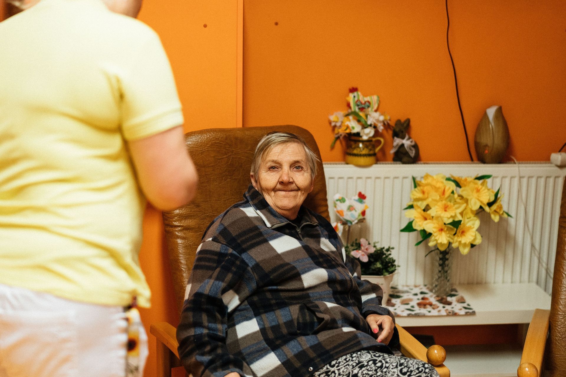 Ältere Frau sitzt lächelnd in einem Sessel und spricht mit einer vor ihr stehenden Person in einem w