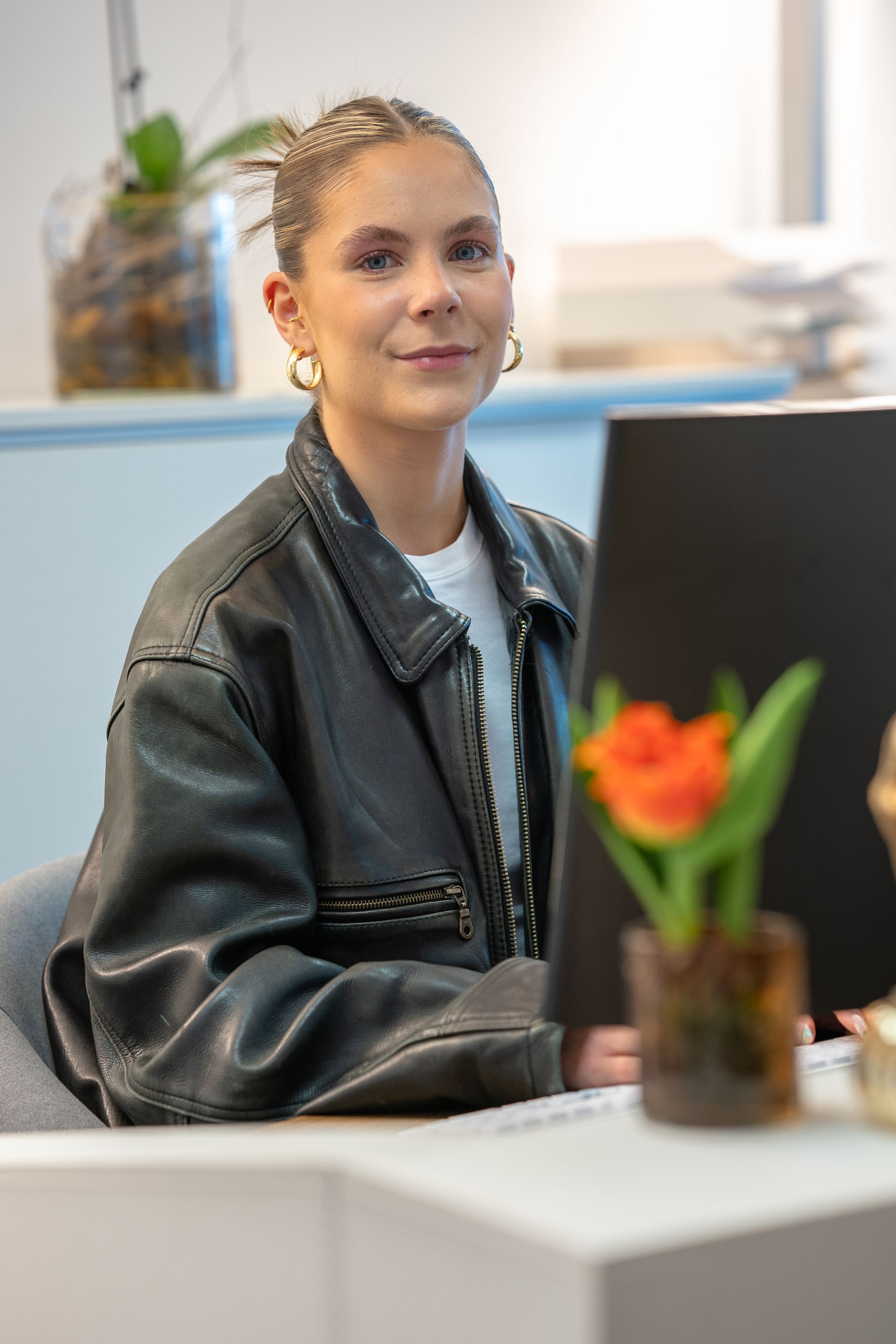 Eine Frau in einer Lederjacke sitzt an einem Schreibtisch vor einem Computer.