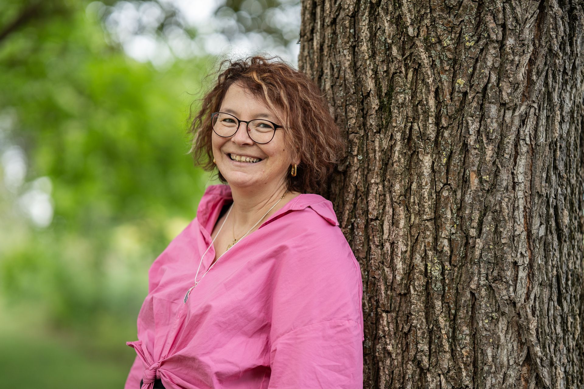 Eine Frau in einem rosa Hemd lehnt an einem Baum und lächelt.