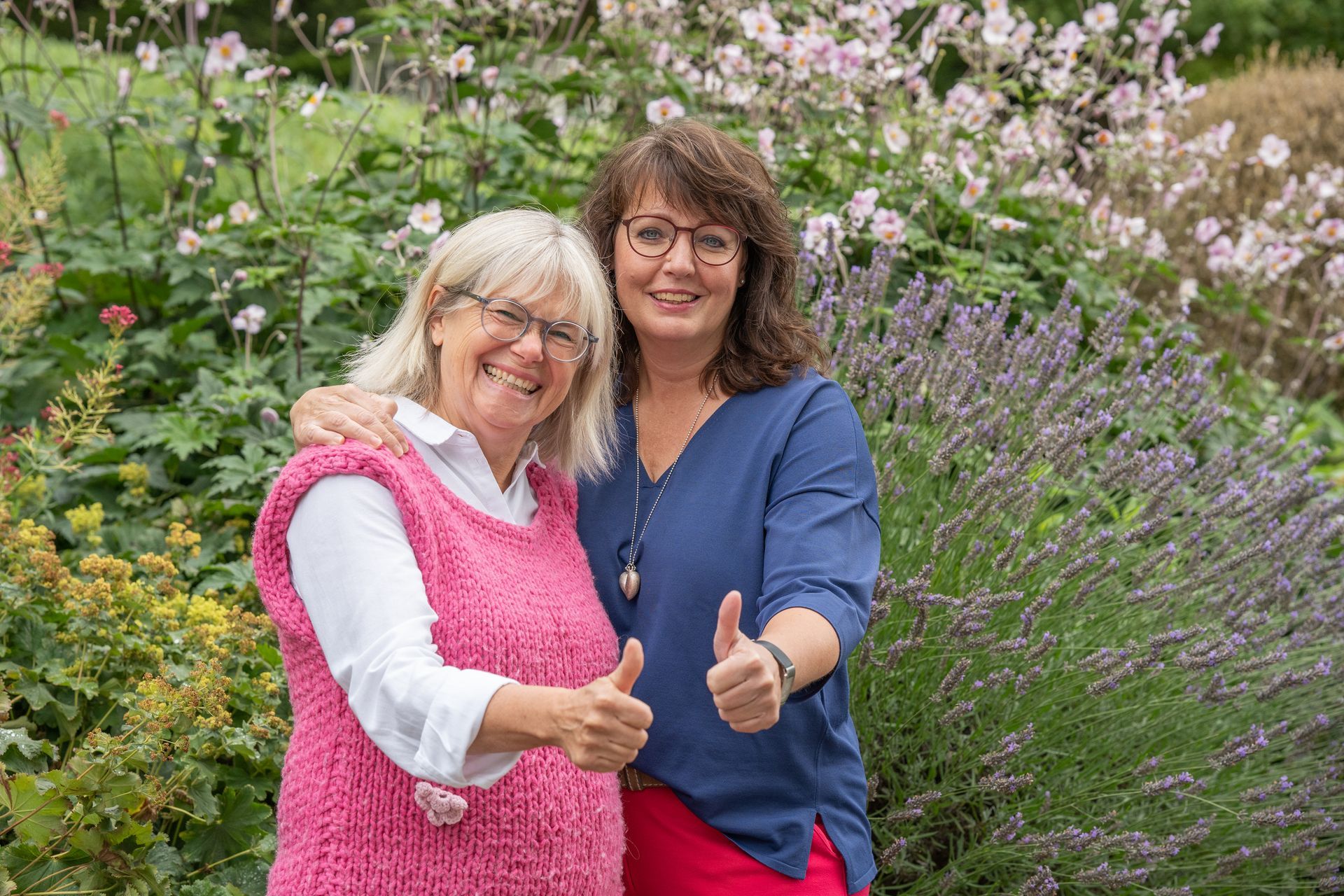 Zwei Frauen stehen nebeneinander vor einem Blumengarten und strecken den Daumen nach oben.
