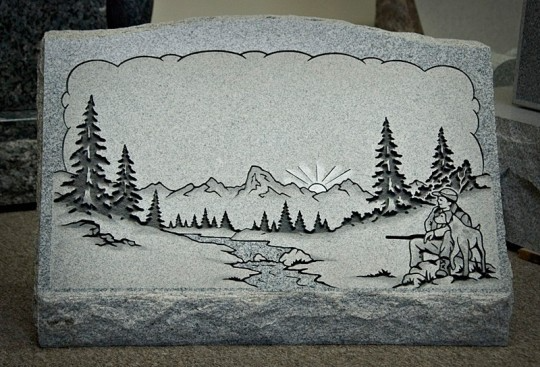 Gravestone with engraved mountain landscape, trees, hunter with dog, and rising sun.
