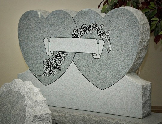 Gray granite headstone shaped like two intertwined hearts, with floral design and blank banner.