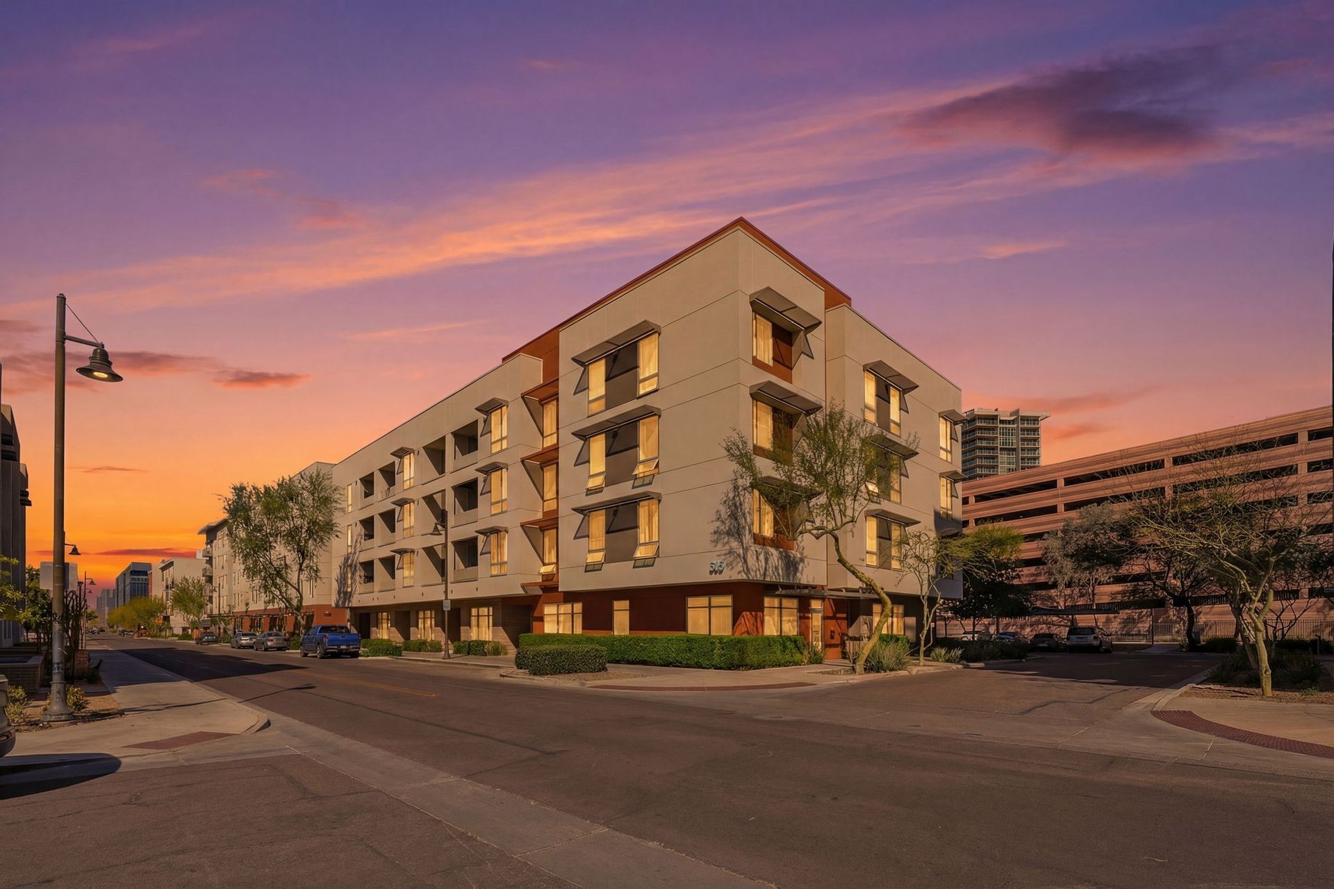 Sunset over a modern apartment building on a quiet street, with pink-purple sky and city lights.