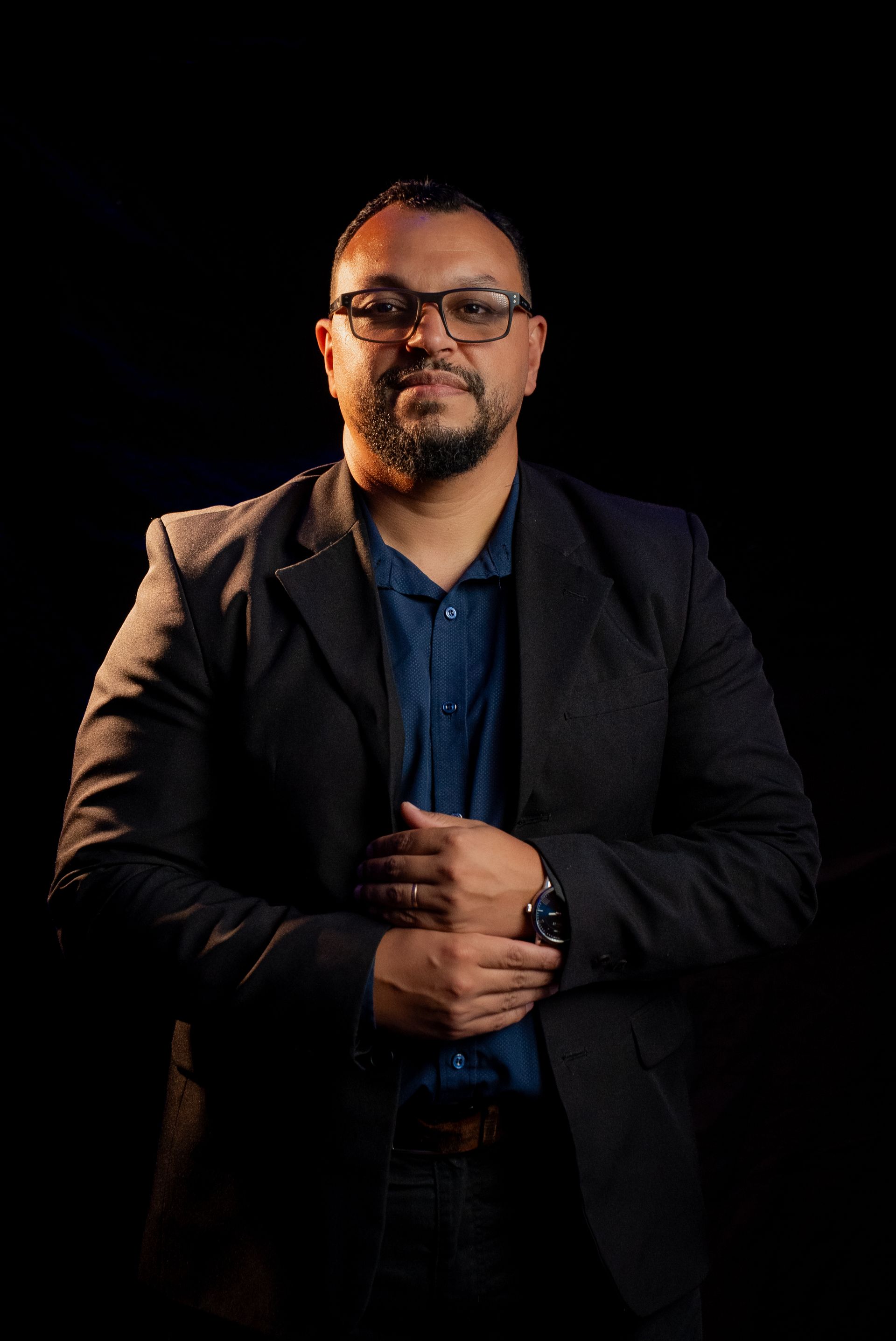 Man in blazer, glasses, hands clasped over stomach, blue shirt. Black background.