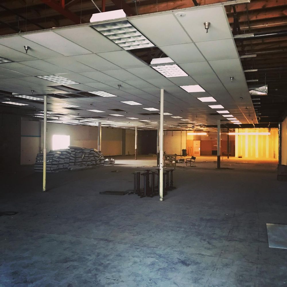 Empty unfinished office with drop ceiling, carpet, tables, stacked materials, and bright sunlight at the far wall