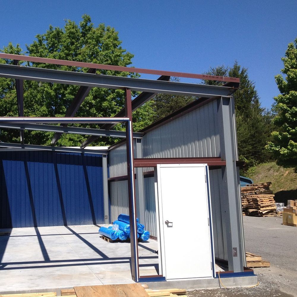 Partially built metal carport beside a small gray shed with a white door, on a sunny day.