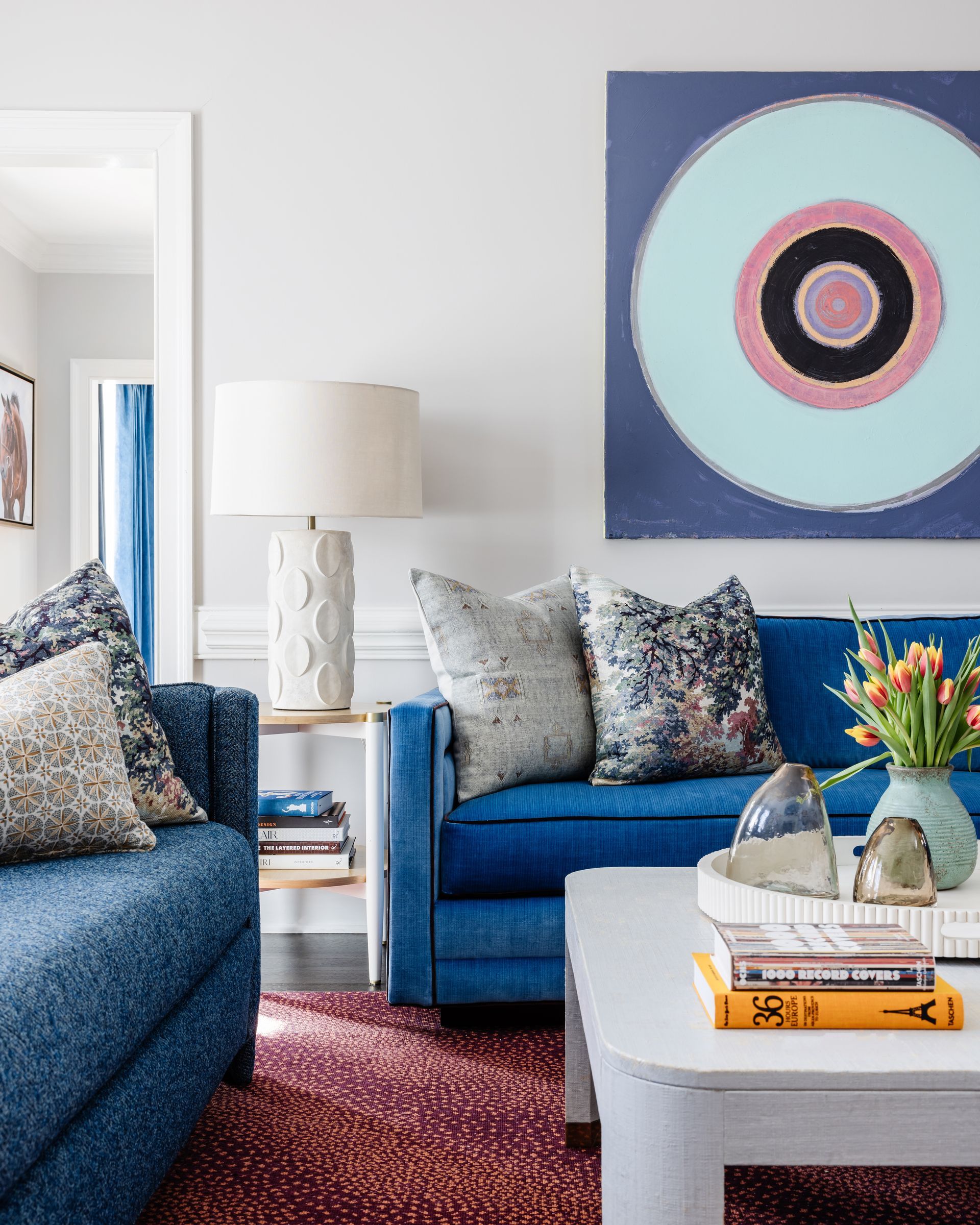 A living room with a blue couch , chair , coffee table and painting on the wall.