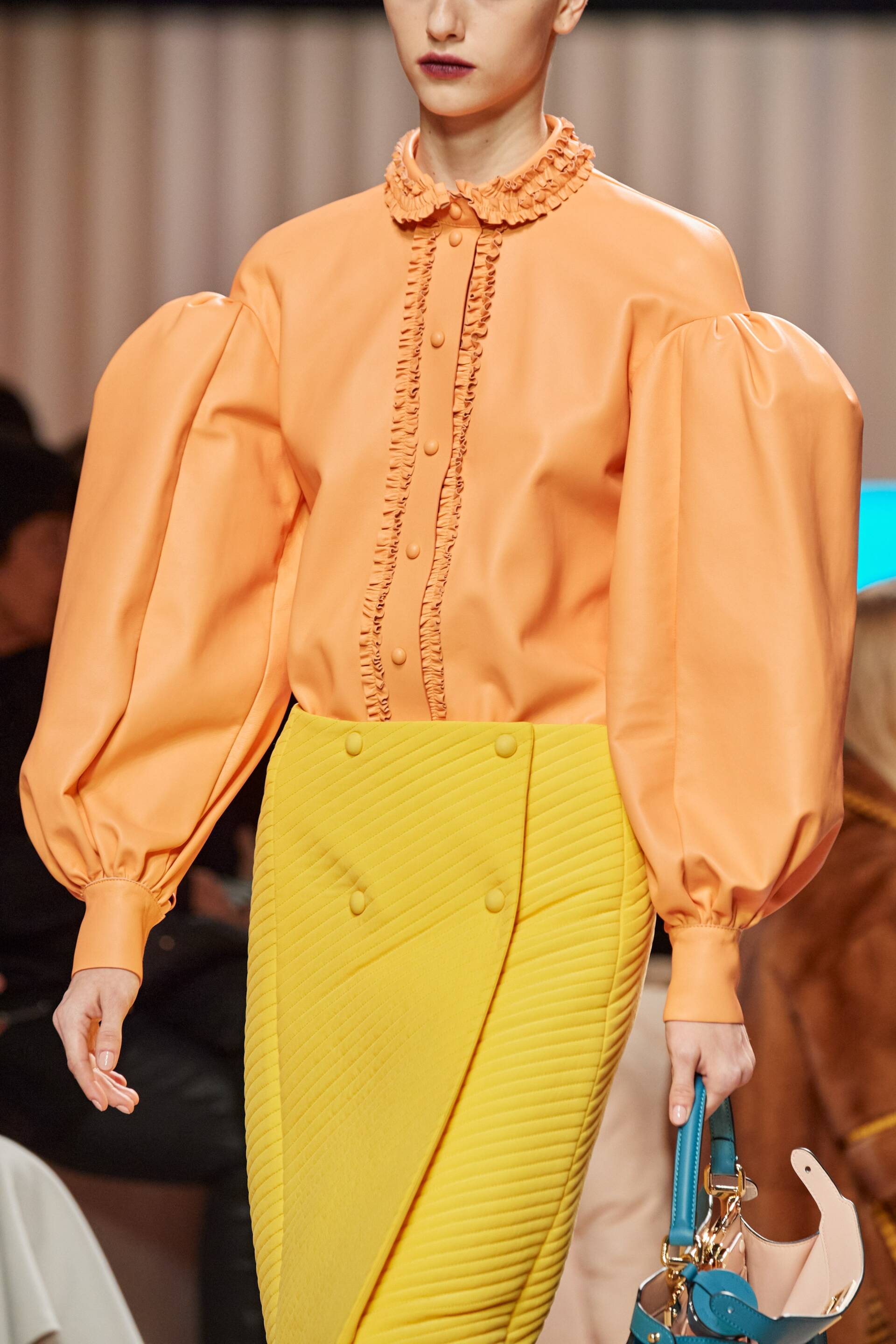 A model wearing an orange shirt and yellow skirt is walking down the runway at a fashion show.