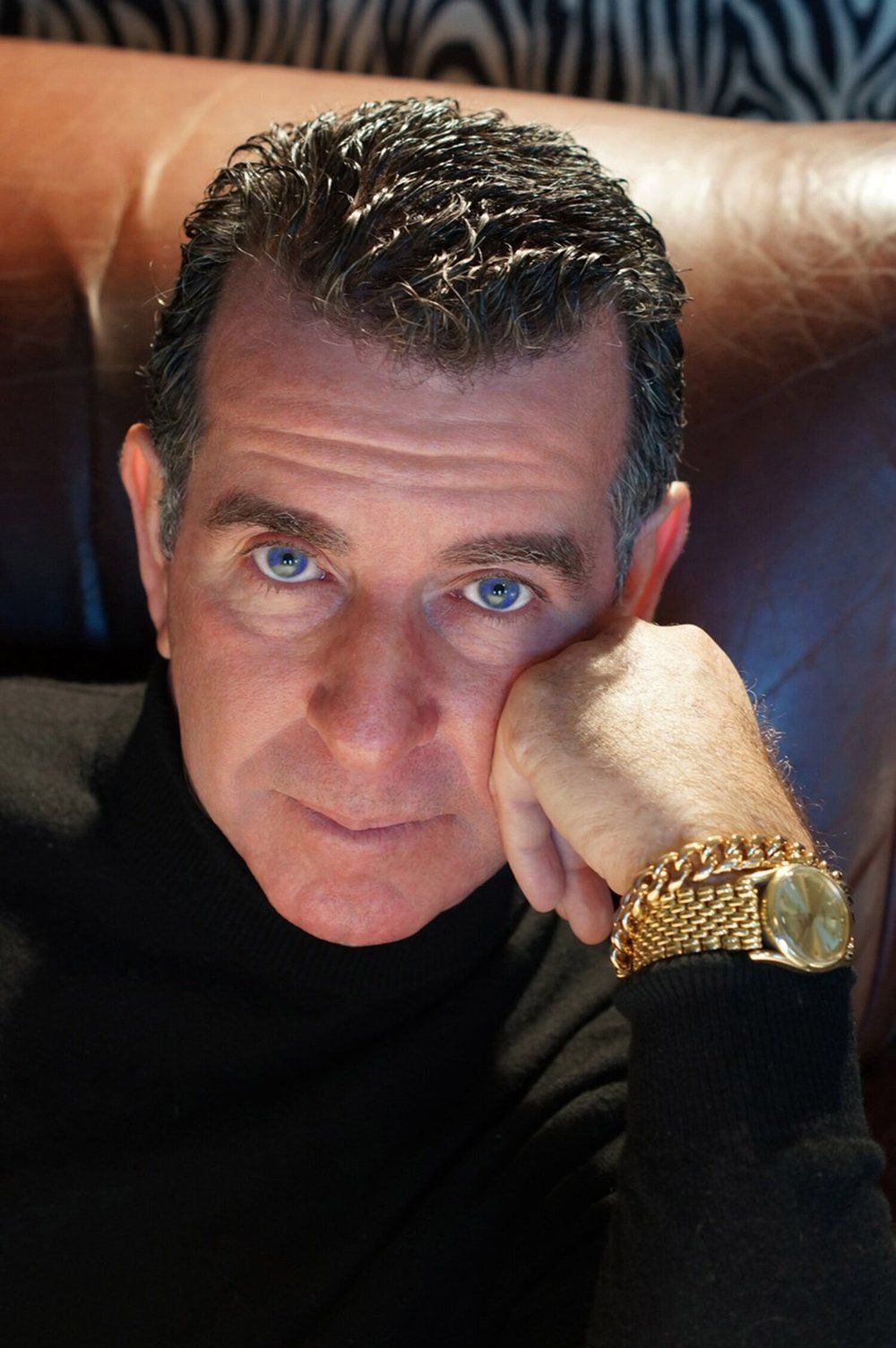 A man wearing a black turtleneck and a gold watch rests his head on his hand