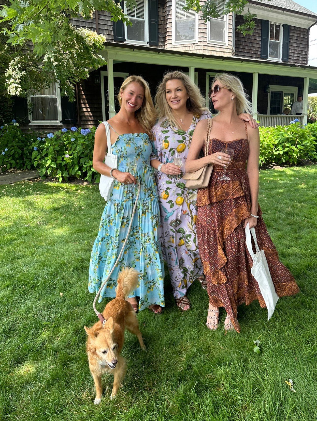 Three women are standing in the grass with a dog in front of a house.