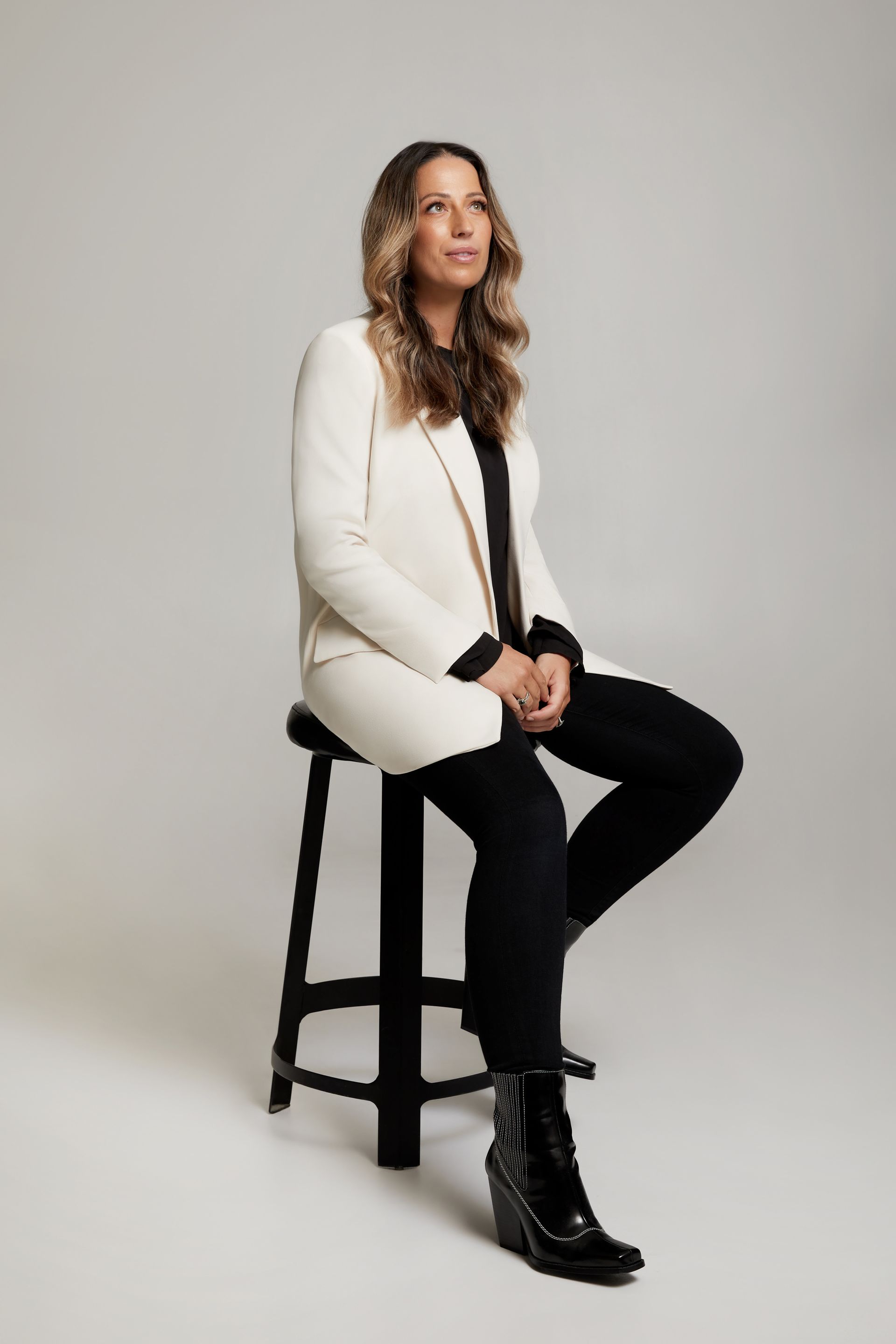 A woman is sitting on a stool wearing a white jacket and black pants.