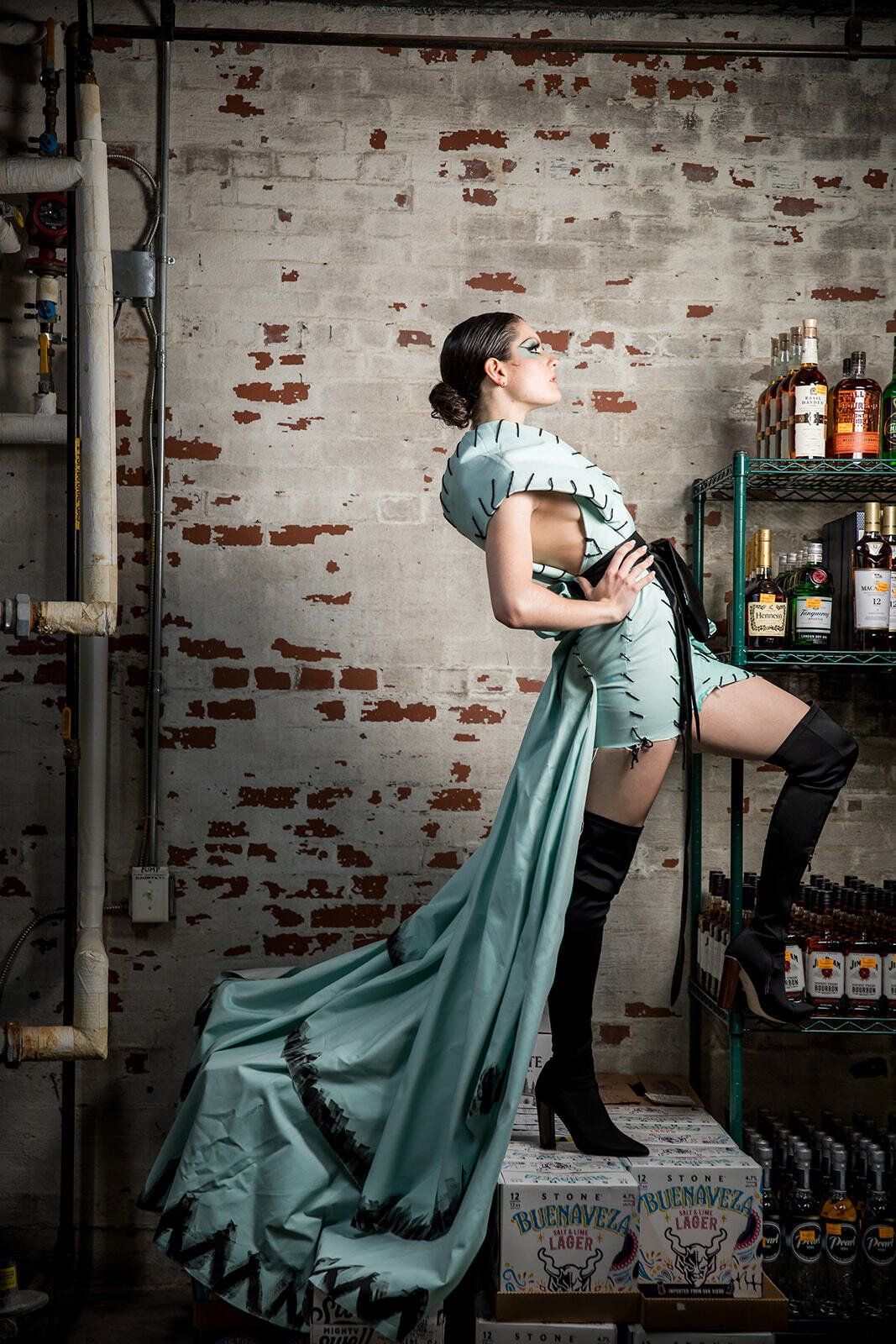 A woman in a long blue dress is standing on a box in front of a brick wall.