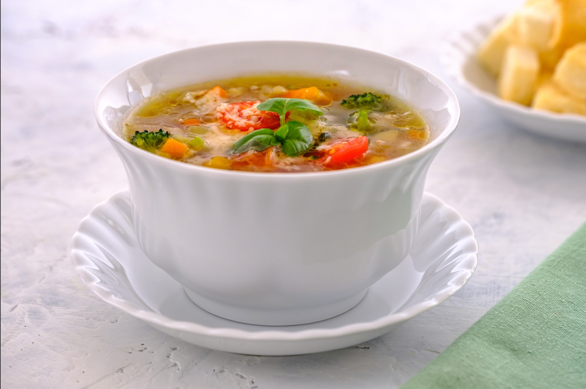 A bowl of soup with broccoli and tomatoes on a saucer on a table.