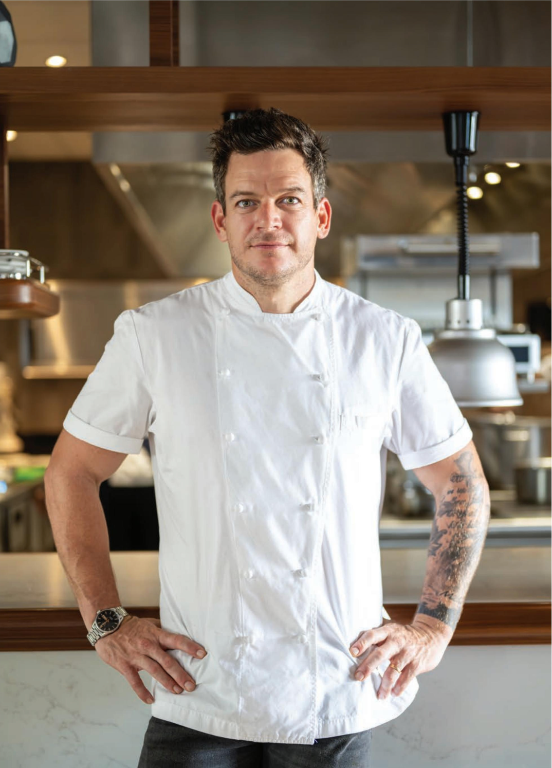 A man in a white chef 's jacket is standing in a kitchen with his hands on his hips.