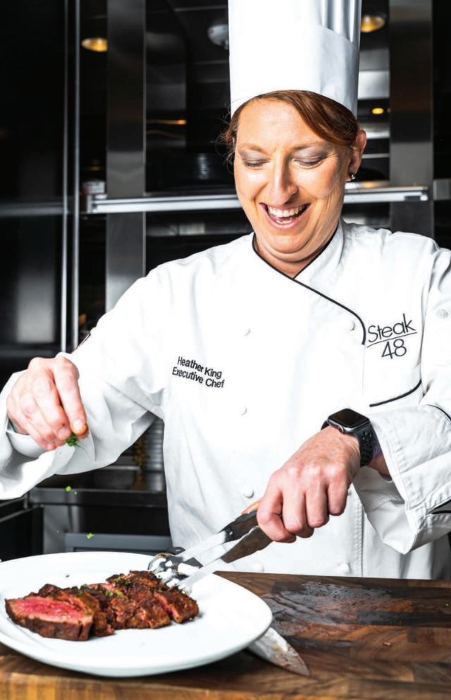 A woman in a chef 's uniform is cutting a steak on a plate.