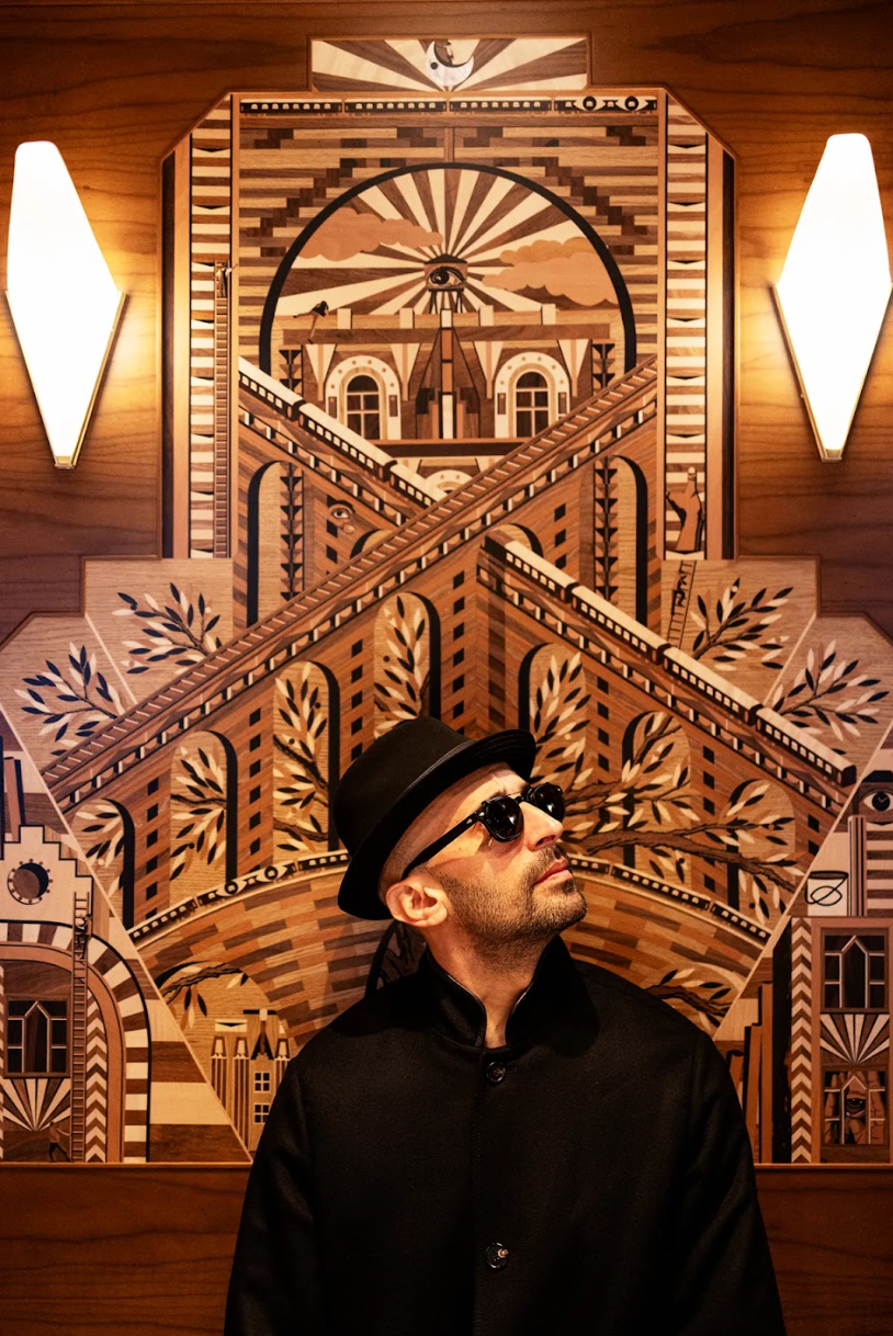 A man wearing a hat and sunglasses is standing in front of a wooden sculpture.