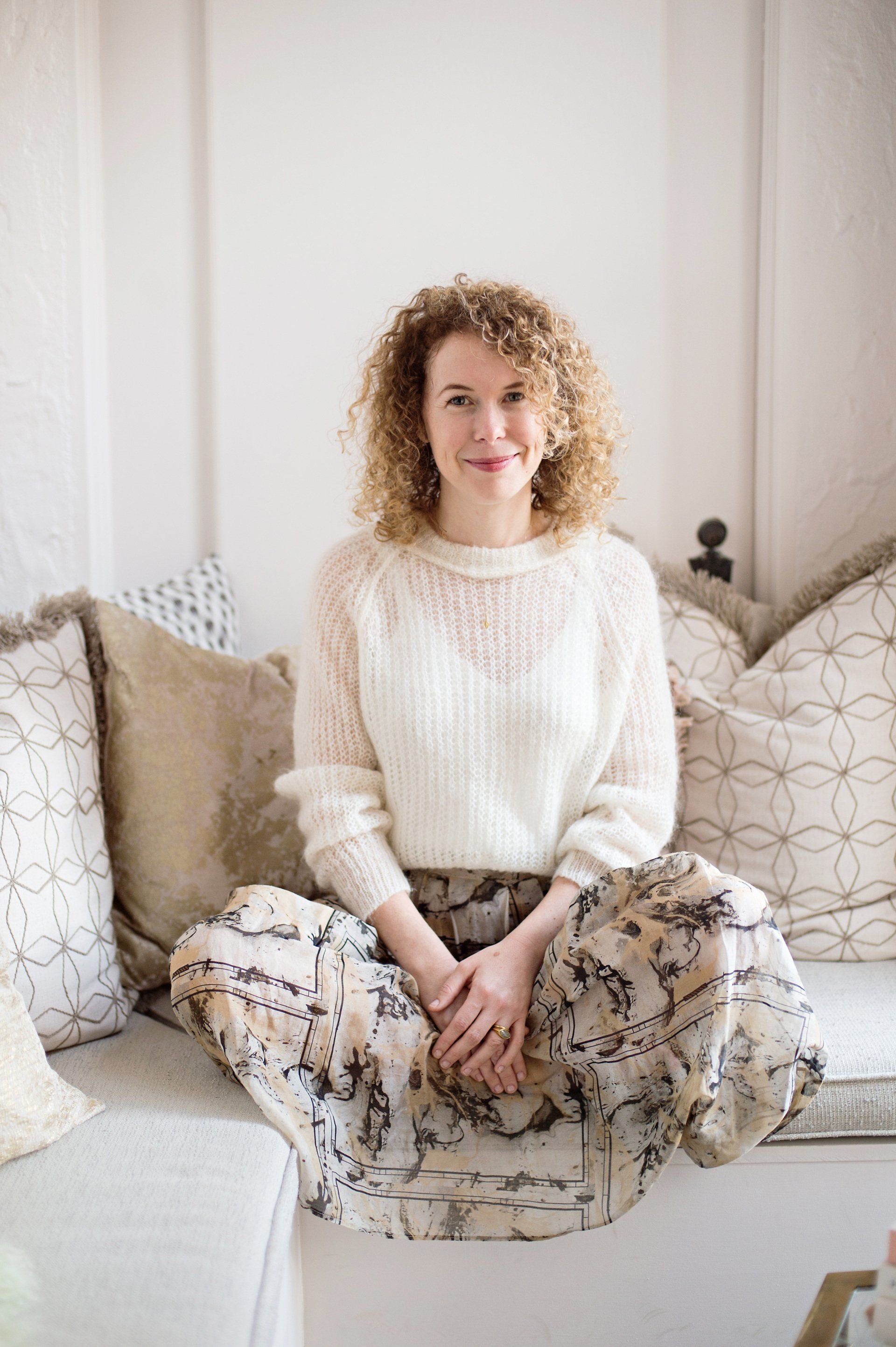 A woman is sitting on a couch wearing a white sweater and a floral skirt.