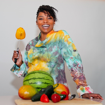 A woman in a tie dye shirt is standing in front of a table full of fruits and vegetables