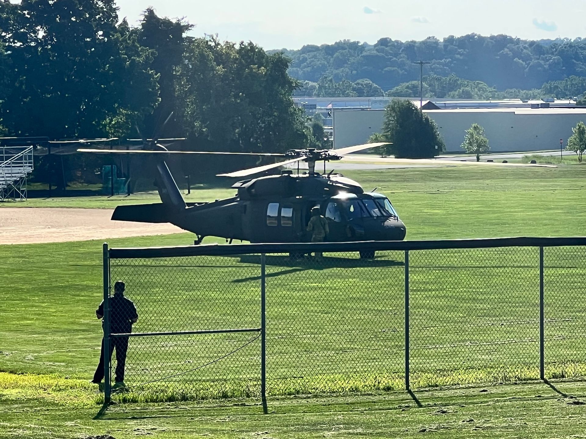 A military helicopter is parked in a grassy field behind a chain link fence.