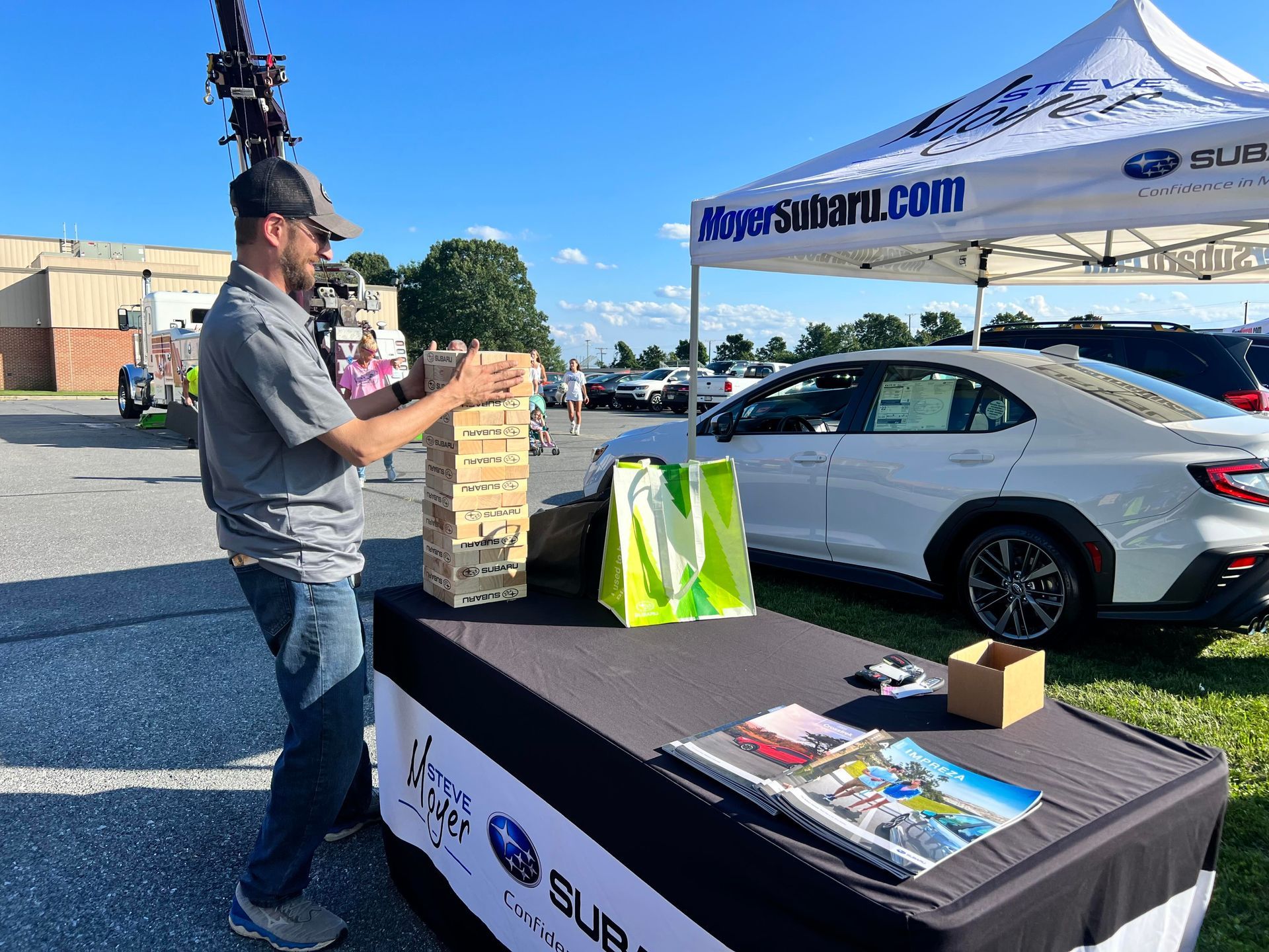 A man is playing jenga at a table in front of a subaru car.