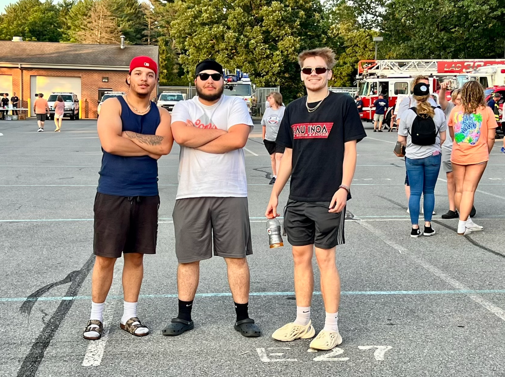 Three young men are standing in a parking lot with their arms crossed.