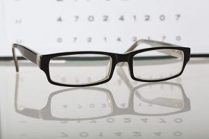 Black-framed eyeglasses on a reflective surface, with an eye chart in the background.