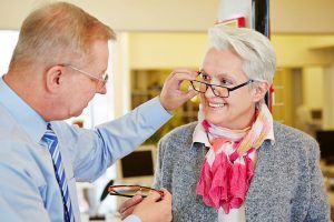 Optician helping an older woman try on eyeglasses in an eye care clinic.