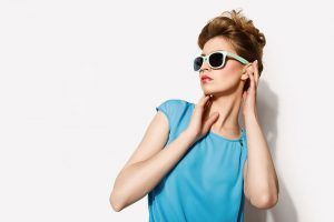 Woman with updo hairstyle wearing sunglasses and blue dress, posing against a white wall.