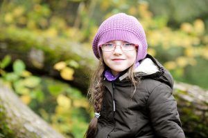 Girl in purple hat and glasses smiles outdoors in front of fall foliage.