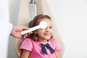 A child at an eye doctor's, smiling as the doctor covers her eye with a white paddle.