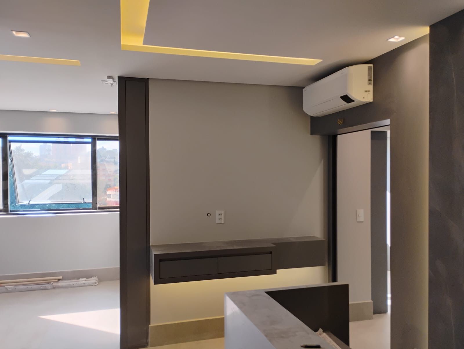 Quarto moderno e vazio com paredes cinza, janela, prateleira fixada na parede e unidade de ar condicionado no teto.