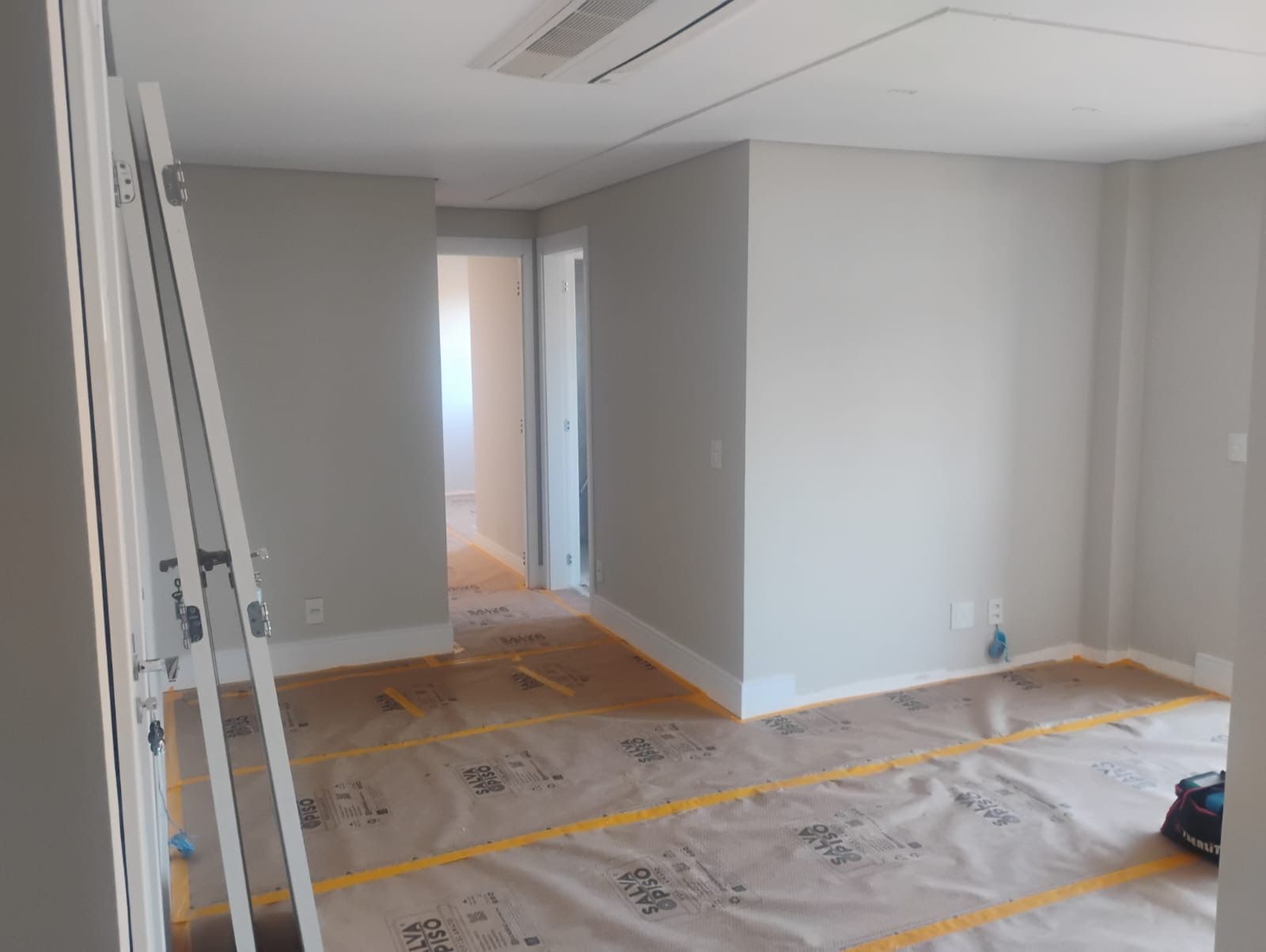 Sala vazia com paredes brancas, piso de madeira e materiais de pintura cobertos por lona plástica.
