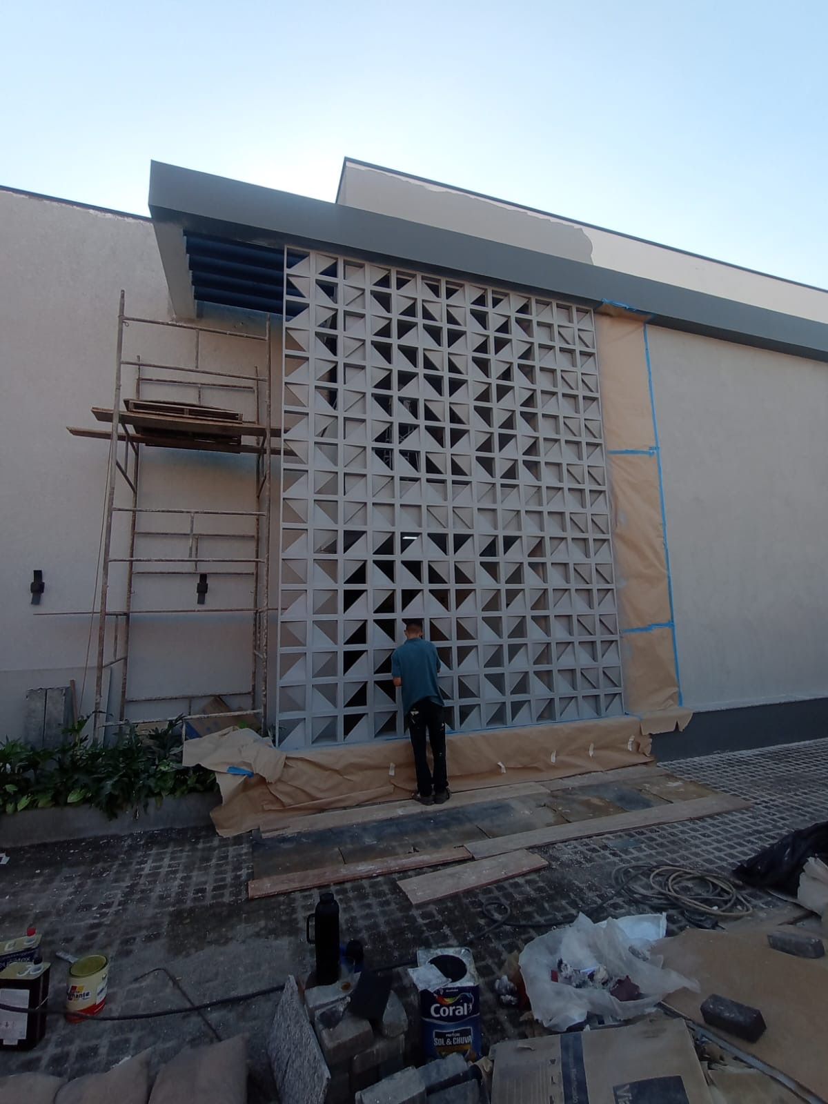 Operário instalando um painel decorativo de concreto na fachada de um prédio, com andaimes e materiais de construção por perto.