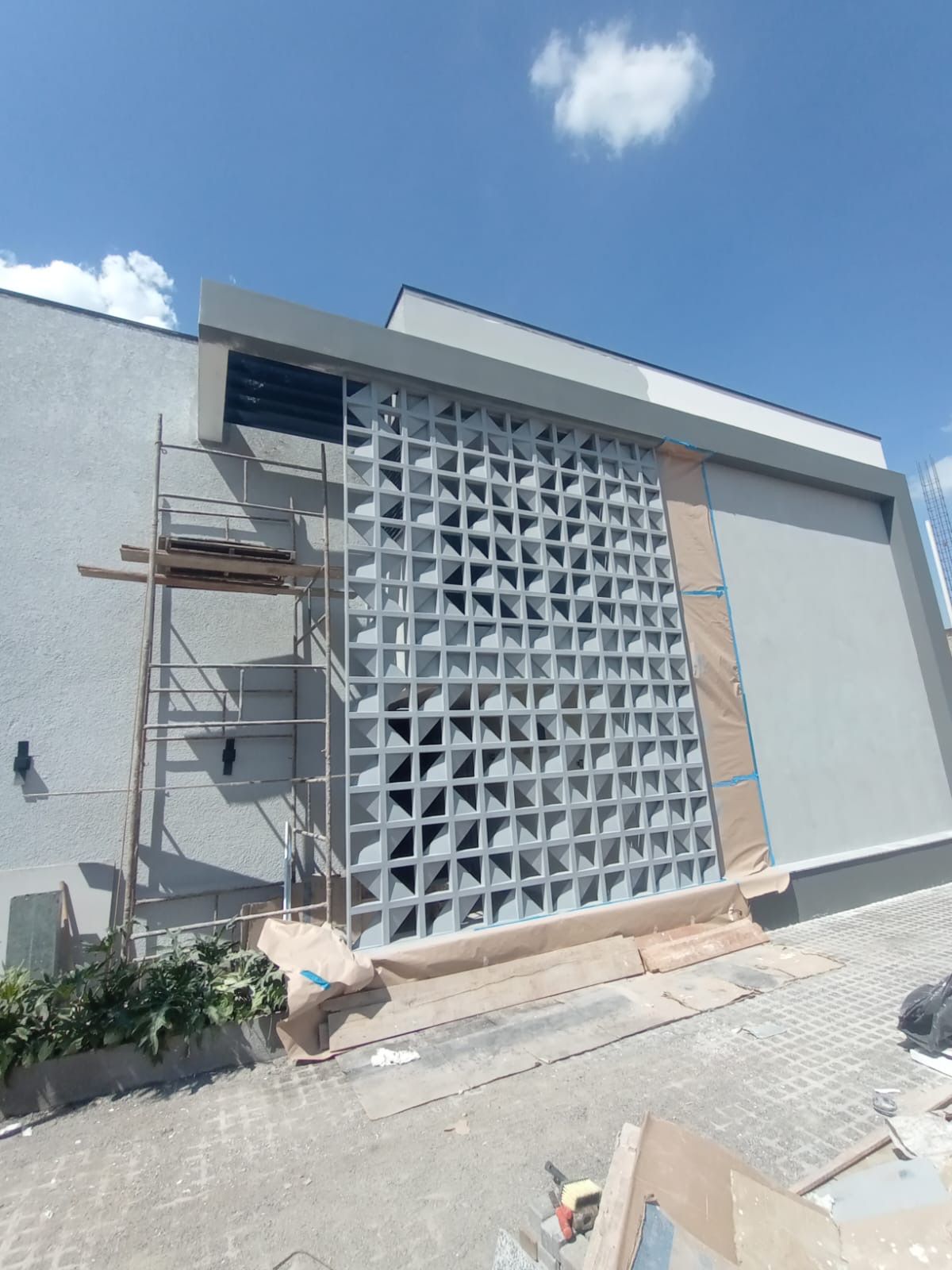 Parede externa com uma grande tela decorativa de blocos de concreto e andaimes sob um céu azul.