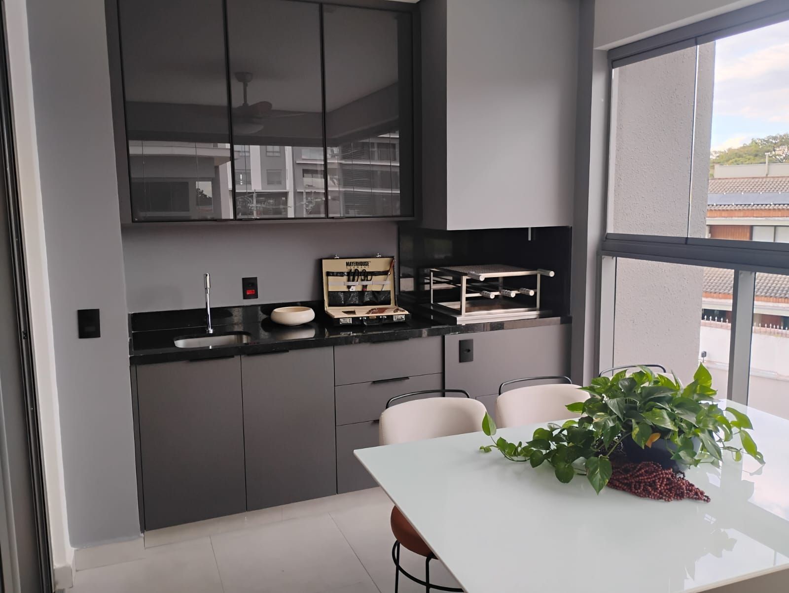 Cozinha moderna com armários escuros, mesa branca e vaso de planta junto a uma grande janela.