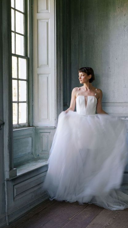 A woman in a wedding dress is standing in front of a window.