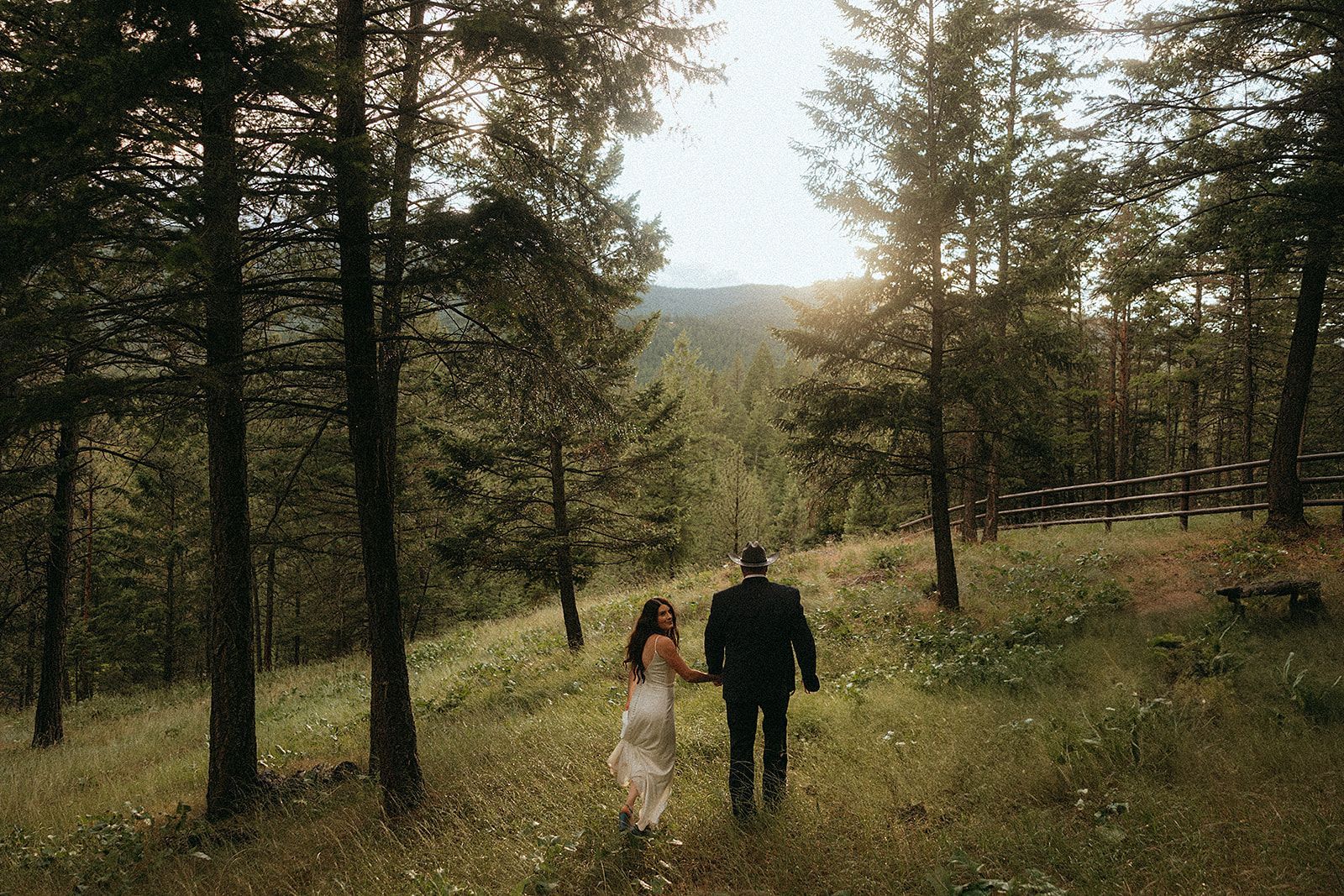 A bride and groom are walking through a forest holding hands.