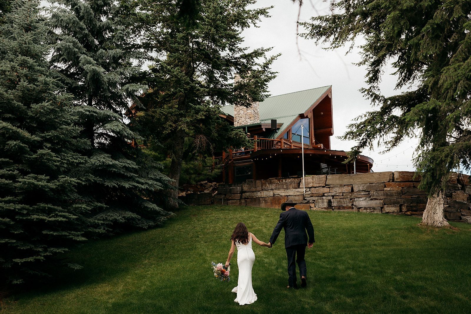 A bride and groom are walking in a grassy field in front of a cabin.